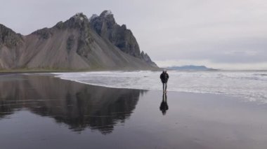 Sırt çantalı adam Vestrahorn Dağı 'ndan ıslak plajda yürüyor.