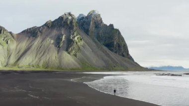 İnsansız hava aracı Vestrahorn Dağı, İzlanda 'nın altından gelen dalgalarla Kara Kum Sahilinde Yürüyen Adam' ın