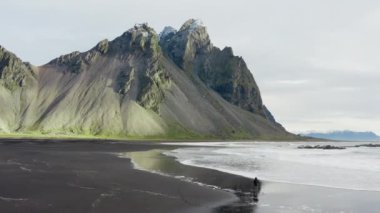 İnsansız hava aracı Vestrahorn Dağı 'na doğru gelen dalgalarla Kara Kum Sahilinde Yürüyen Adam' ın görüntüsü.