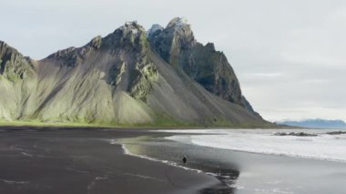 İnsansız hava aracı Vestrahorn Dağı, İzlanda 'nın altından gelen dalgalarla Kara Kum Sahilinde Yürüyen Adam' ın