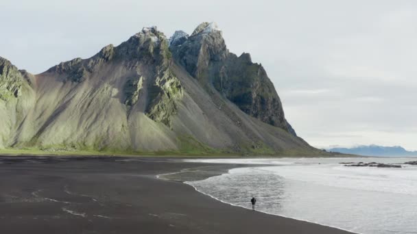 アイスランドのヴェストラホーン山の下に来る潮と黒砂のビーチを歩く男のドローンショット — ストック動画