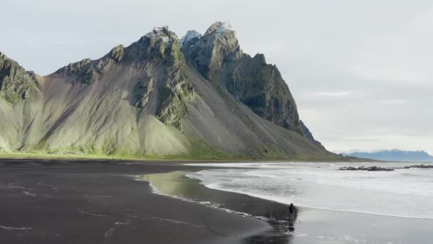 アイスランドのヴェストラホーン山に向かって来る潮と黒砂のビーチを歩く男のドローンショット — ストック動画