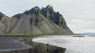 Siyah kumsalda yürüyen ve dalgalar boyunca koşan bir adamın robot resmi. Arka planda Vestrahorn Dağı, İzlanda.