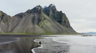Siyah kumsalda yürüyen bir adamın Drone 'u dalgalar içeri girerken ve arka planda Vestrahorn Dağı, İzlanda manzarasına bakmak için durduğunda çekildi.
