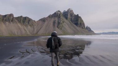 İzlanda 'daki Vestrahorn Dağı' na doğru siyah kum plajı boyunca sırt çantasıyla yürüyen üç ayaklı yürüyüşçünün Statik Yavaş Hareketi.