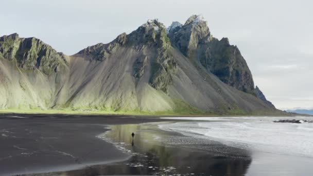 アイスランドのヴェストラホーン山に向かって来る潮で黒い砂のビーチを歩く男のドローンショットをアーチ — ストック動画