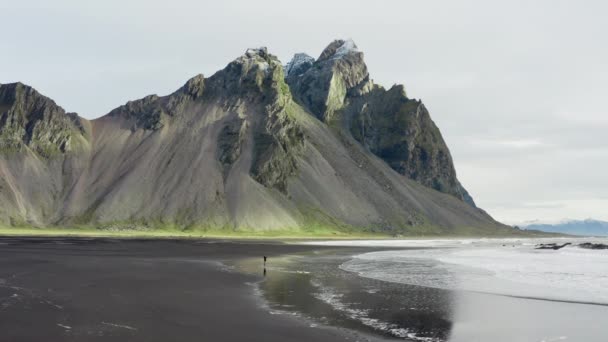 彼はヴェストラホーン山に向かって実行されるように潮が入ってくる黒砂のビーチで男のドローンショット アイスランド — ストック動画