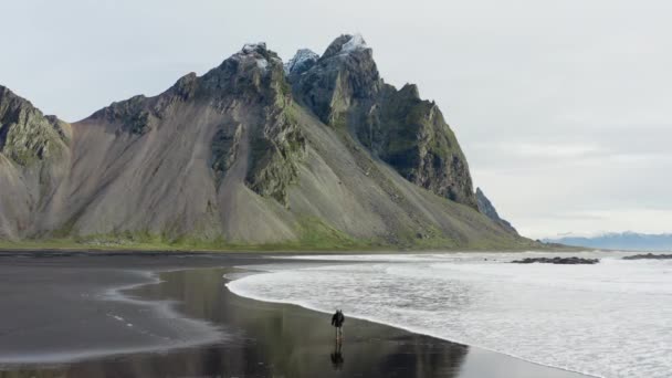 黒砂のビーチで男のドローンショット歩くと背景にヴェストラホーン山と来る潮に沿って実行 アイスランド — ストック動画