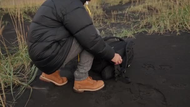 Muž rozepínání batoh na černé písečné pláži — Stock video
