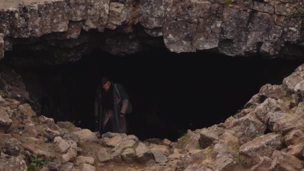 Людина в довгому пальто з гітарним футляром залишаючи печеру — стокове відео