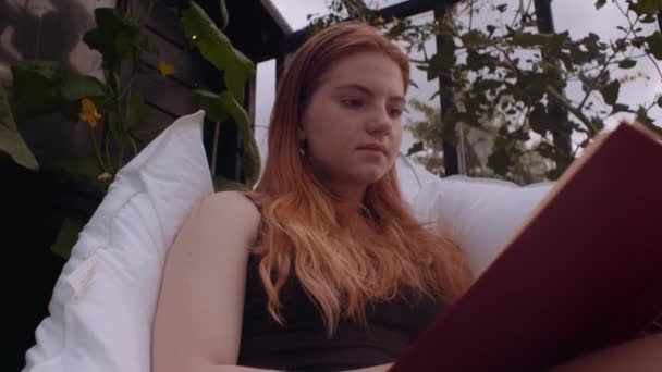 Adolescente assise dehors sur des oreillers dans le jardin et le livre de lecture — Video