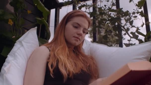 Tenåringsjente sitter utenfor på puter i hagen og leser bok – stockvideo