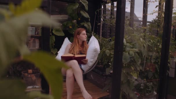 Tonårstjej sitter på trädgårdsstol med kuddar och läser — Stockvideo