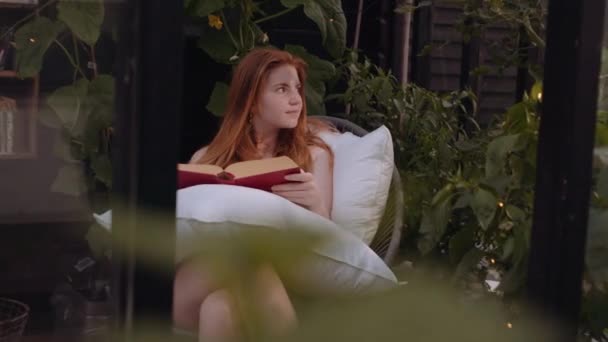 Adolescente chica sentado fuera en almohadas en jardín y lectura libro — Vídeos de Stock