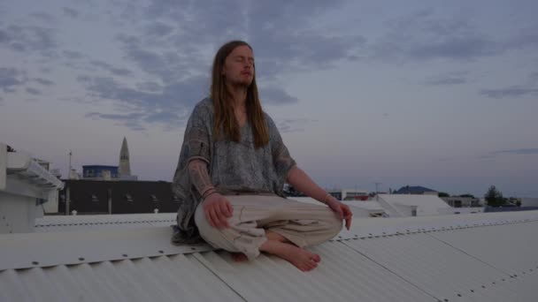 Ung man med långt hår mediterar på stadens tak — Stockvideo