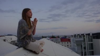 Uzun saçlı genç adam şehrin çatısında meditasyon yapıyor.