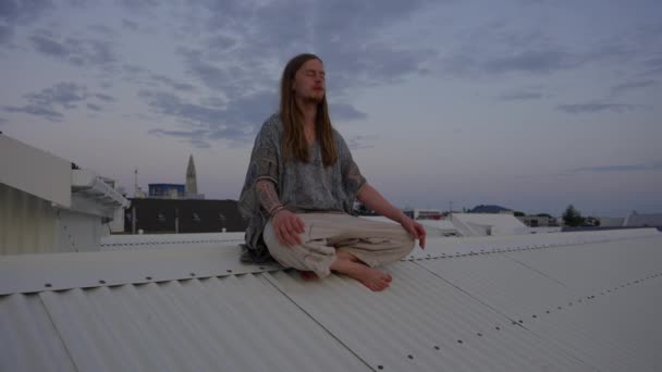 Jeune homme aux cheveux longs méditant sur le toit de la ville — Video