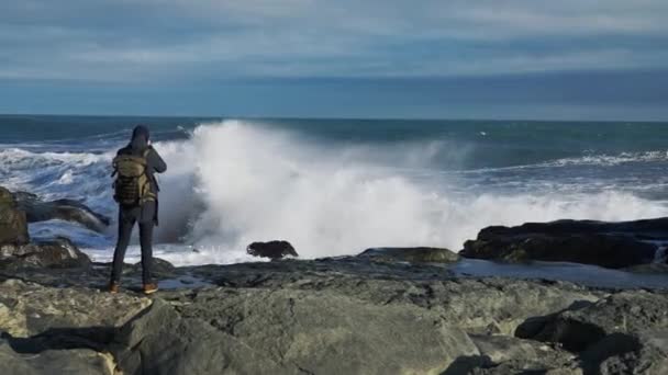 Человек с рюкзаком, стоящим на скалах, фотографирует волны — стоковое видео