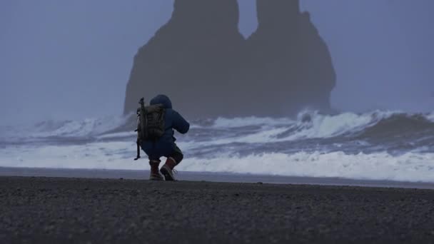 Fotograf przyczajony na czarnej plaży z burzliwym morzem — Wideo stockowe
