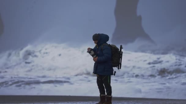 Фотограф Чистящий объектив на Черном песке с бурным морем — стоковое видео