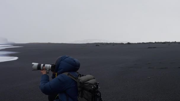 Fotógrafo sosteniendo la cámara para fotografiar desde la playa de arena negra — Vídeos de Stock