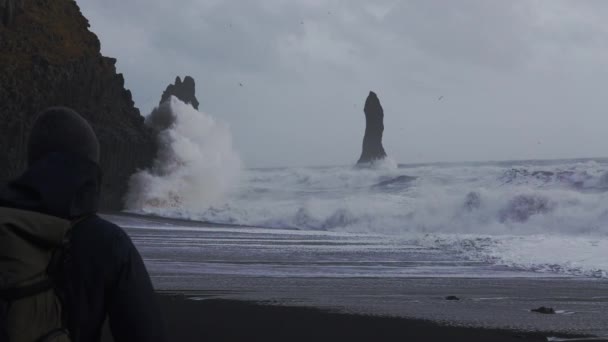 Fotógrafo caminando sobre la playa de arena negra hacia el surf blanco — Vídeos de Stock