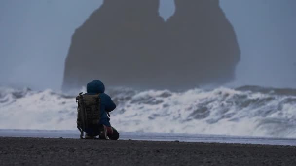 Fotógrafo agachándose para fotografiar el mar áspero de la playa de arena negra — Vídeos de Stock