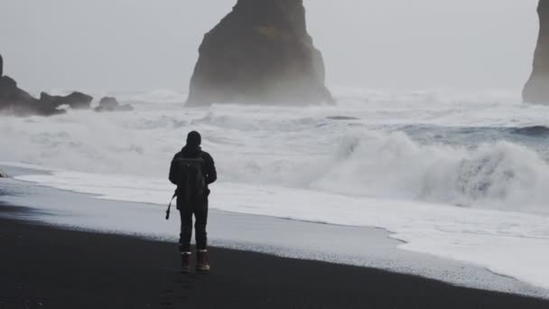 Fotógrafo caminando a lo largo de la playa de arena negra para fotografiar mar áspero — Vídeos de Stock