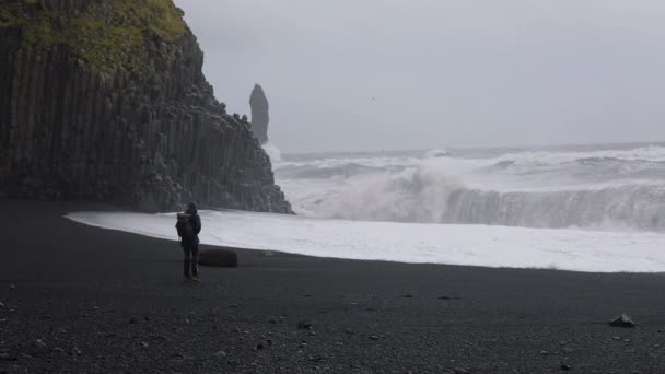 Fotógrafo parado en la playa de arena negra frente al mar áspero y tormentoso — Vídeos de Stock