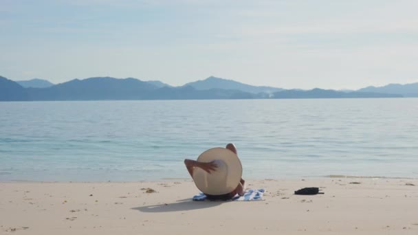 Kobieta w kostiumie kąpielowym i kapeluszu wstaje, by usiąść i opalać się na plaży. — Wideo stockowe