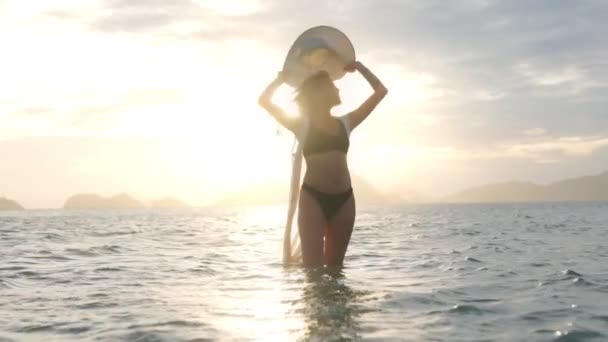 Žena v bikinách se slunečním kloboukem, stojící v moři při západu slunce — Stock video