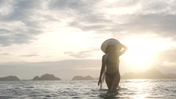 Vrouw in bikini en zonnehoed wandelen in zee als de zon ondergaat — Stockvideo