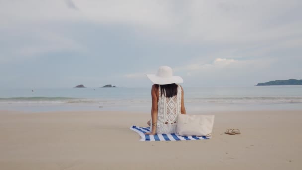 Frau im Sonnenhut sitzt auf Strandtuch und blickt aufs Meer — Stockvideo