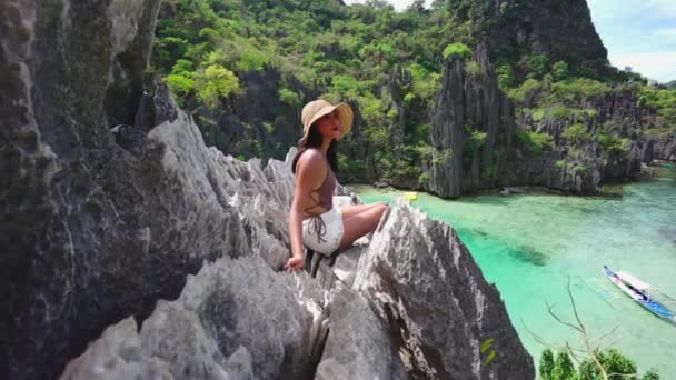 Женщина в солнечной шляпе сидит на скалах над скрытым пляжем — стоковое видео