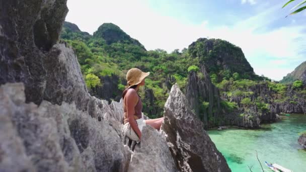 Женщина в солнечной шляпе сидит на скалах над скрытым пляжем — стоковое видео