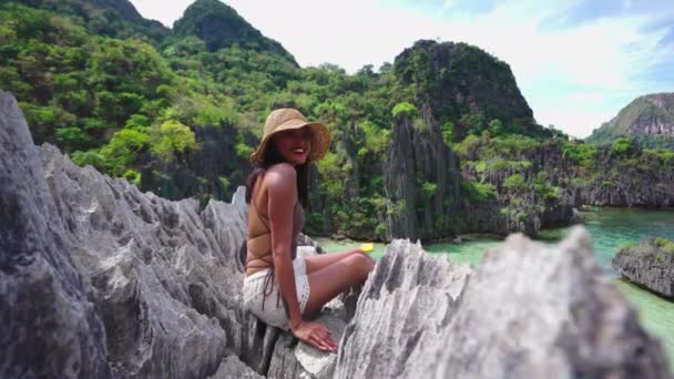 Mujer sonriente con sombrero de sol sentada en rocas sobre la playa oculta — Vídeos de Stock