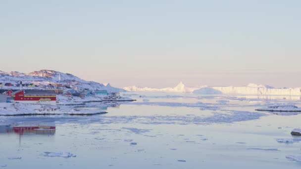 広いスローモーションは グリーンランドのイルリサットの氷と海に向かって海岸線を越えて太陽の光が当たる町から出てくる — ストック動画