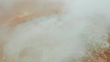 Myvatn Gölü yakınlarındaki Hver Jeotermal Bölgesi 'nde Buharlı Fumarollerin Önünde İki Turistin Hava Çekimi. Geniş açılı panoramik çekim. Kamera ileri doğru hareket ediyor. Drone Buharda Uçuyor