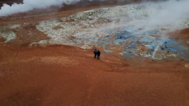 2人の観光客の空中ショットMyvatn湖の近くのHverir地熱地域の蒸しフマロールの前に立っています パノラマ高角度ワイド軌道ショット — ストック動画