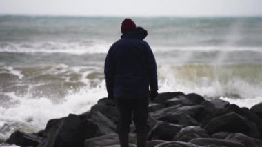 Kışlık Kıyafetli Adam, Rocky Sahili 'nde Deniz kenarında duruyor ve Gelen Fırtınayı ve Büyük Dalgaların Kıyıya Çarpışmasını izliyor. Yavaş Hareket Görüntüsü