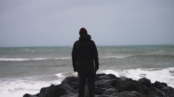 穿着冬衣的男人站在岩石海岸的海边 看着即将到来的风暴和巨浪冲击着海岸 慢动作射击 — 图库视频影像