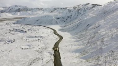 Aerial of Car Driving on a Dirt Road Along the Snow Covered Mountains in Iceland. High Altitude Panoramic Shot. Camera Tracking the Car