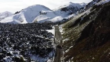 Aerial of Car Driving on a Dirt Road Along the Snow Covered Mountains and Black Lava Fields in Iceland. Panoramic Shot. Camera Tracking the Car