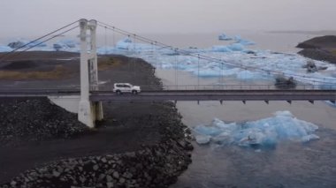 Aerial of a Car Driving on a Cable Bridge Above the Glacial River with Large Ice Splinters, Iceland. Camera Follows the Moving Car