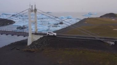 Aerial of a Car Drives Off a Cable Bridge Above the Glacial River  with Large Ice Splinters, Iceland. Aerial Panning Shot. Camera Moves Right