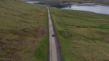 Aerial of a Car Driving Along an Empty Rural Road In Iceland. Car Passes by Majestic Mountains and Green Meadows. Wide High Angle Shot. Camera Tracking the Car