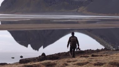 Sırt çantalı ve Kameralı Adam Yüzeyinde Dağların Yansımaları ile Göle Doğru Yürüyor. Arka Görünüm Statik Görüntüsü