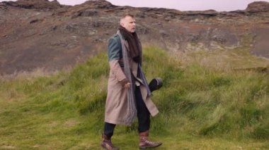 Stylish Man with a Sack Bag on His Shoulder and a Banjo Case in His Hand Walks Towards Little Black House. Cinematic Slow Motion Pan Side View Shot. Camera Tracking the Man