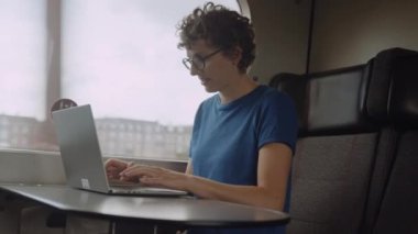 Woman with Curly Hairs and with Glasses On Working on a Laptop While Going by Train. She Is Sitting at the Table by the Window