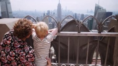 Medium Slow Motion Arcing Shot Over Mother And Young Son Looking Through Railings On Rooftop To New York Skyscrapers